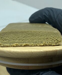 Bloc de résine CBD compact et clair, posé sur une planche en bois, avec une main gantée en arrière-plan.