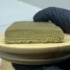 Bloc de résine de cannabis 3x filtré, texture fine et homogène, posé sur un plateau en bois, tenu par une main gantée.