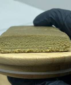 Bloc de résine de cannabis 3x filtré, texture fine et homogène, posé sur un plateau en bois, tenu par une main gantée.